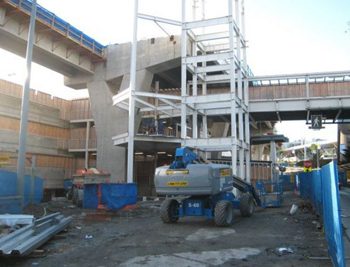Structural steel of the glass-enclosed staircase tower being assembled in the foreground, with the concrete station platform in the background.